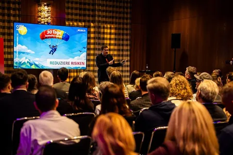 Christian Dietz auf der Bühne beim OMT Summit, vor ihm das Publikum, auf dem Bildschirm die Folie „Reduziere Risiken“