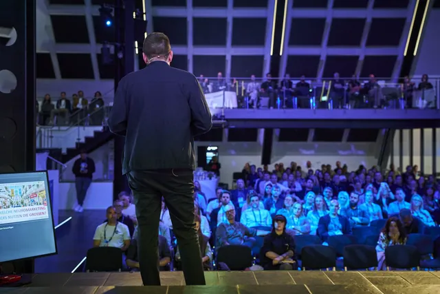 Blick von der Bühne über Christian Dietz hinweg in den vollbesetzten Saal mit Empore beim OMT 2026
