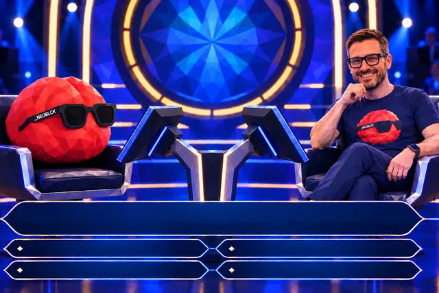 FRED, das rote Gehirn mit NEUBLCK-Brille, und Christian Dietz im FRED-T-Shirt sitzen in einem Fernsehstudio im Stil von „Wer wird Millionär?“ in zwei Sesseln, vor ihnen leere Frage- und Antwortfelder