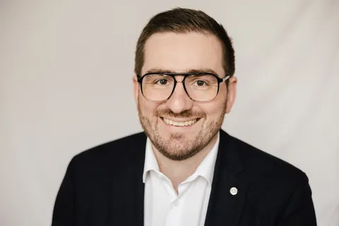 Christian Dietz smiling against a light background, wearing glasses, a dark jacket and a white shirt