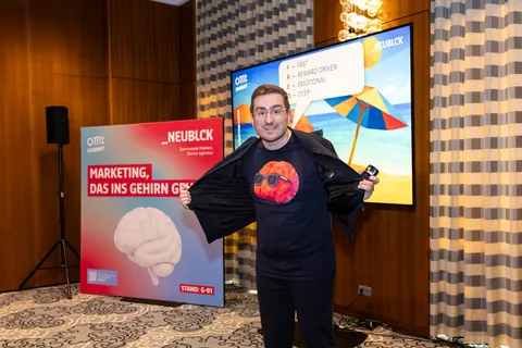 Christian Dietz opens his jacket to show the FRED shirt with the orange brain wearing sunglasses, at the NEUBLCK stand at the OMT Summit