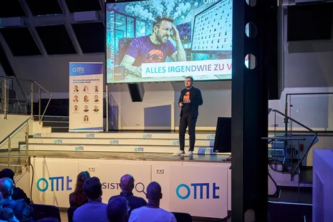 Christian Dietz on stage at OMT 2026 in Mainz, a slide with his picture on the big screen above him, the audience in front